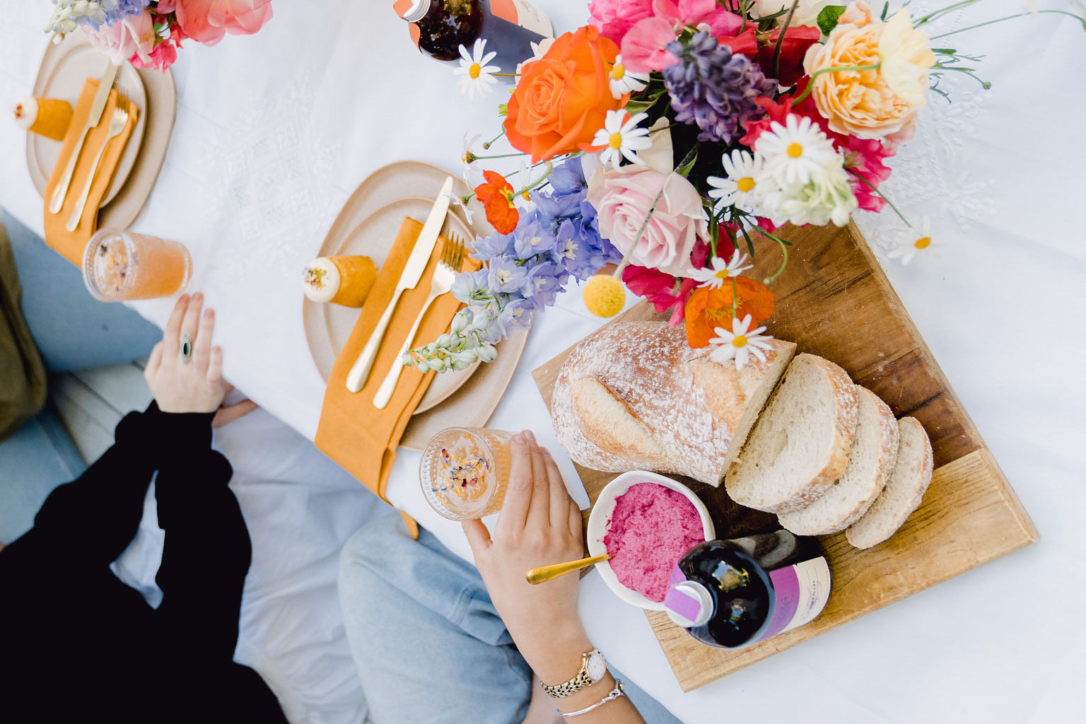 Person enjoying a meal with bread, flowers, and drinks on a table.